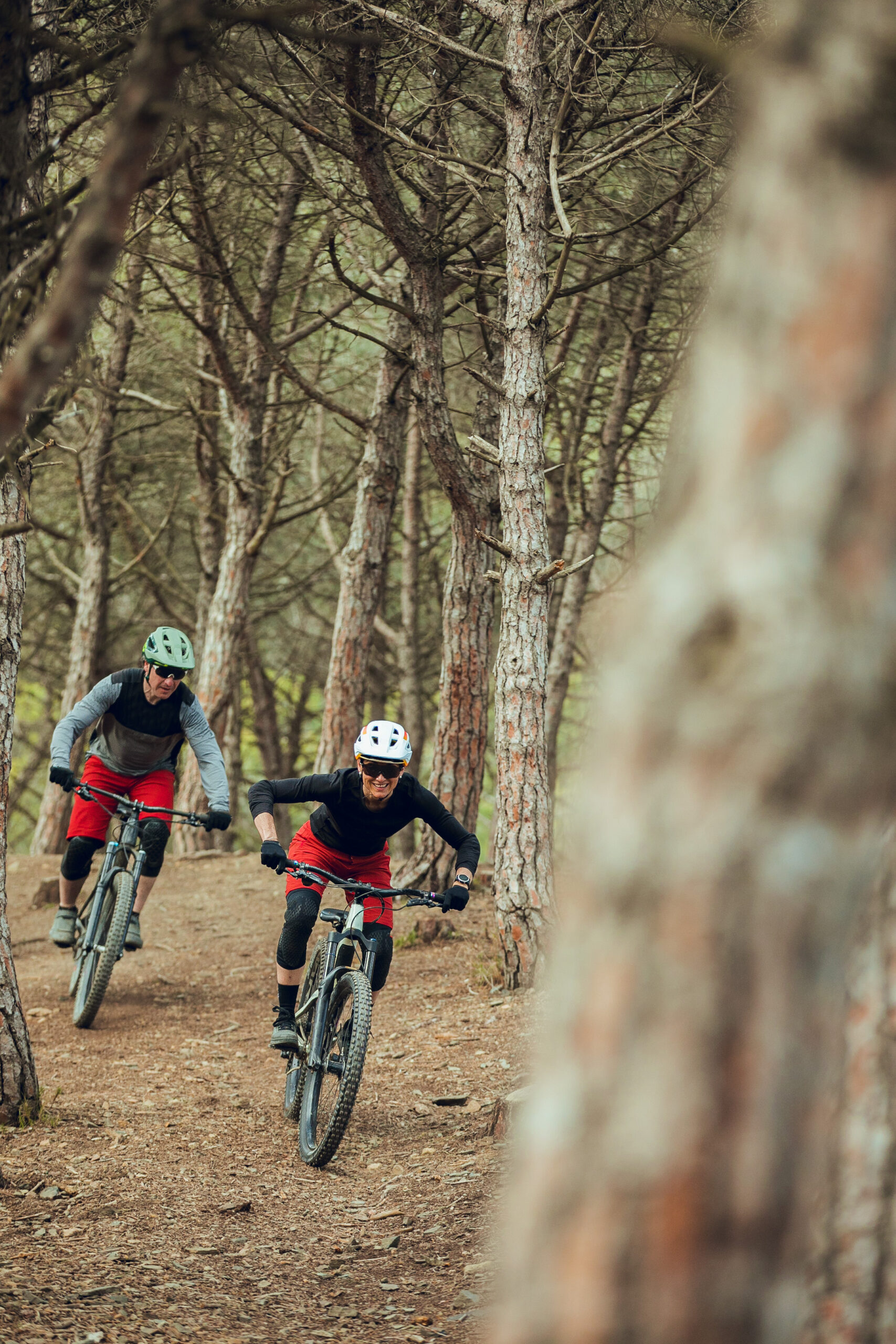 Couple of enduro mtb riders, woman and man dressed in black and red with helmet, gloves and goggles going down the mtb trail in summer in the forest